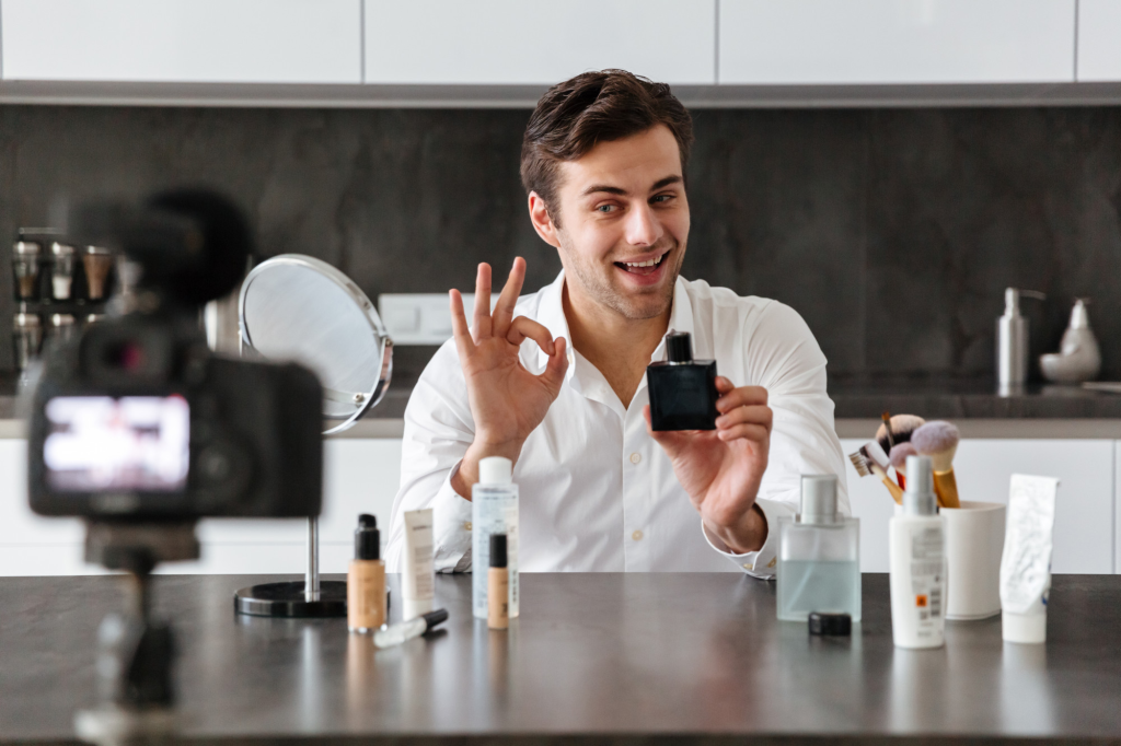 Mand holder parfume og laver OK-tegn mod kameraet.
